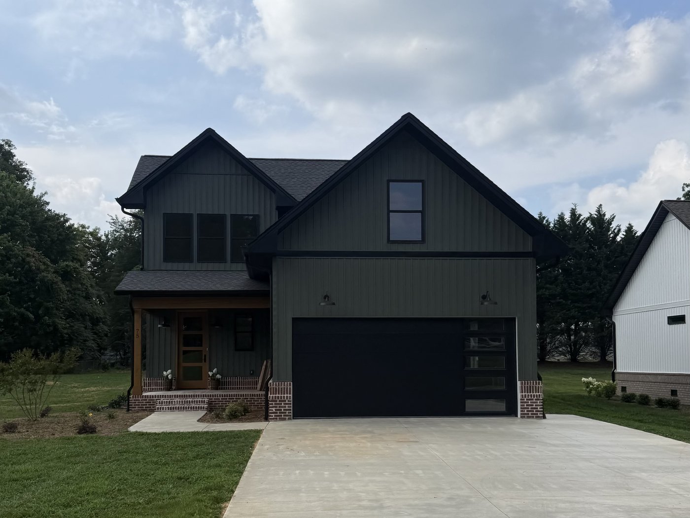 Completed olive-green two-story modern farmhouse with black garage door, brick skirt, and finished concrete driveway